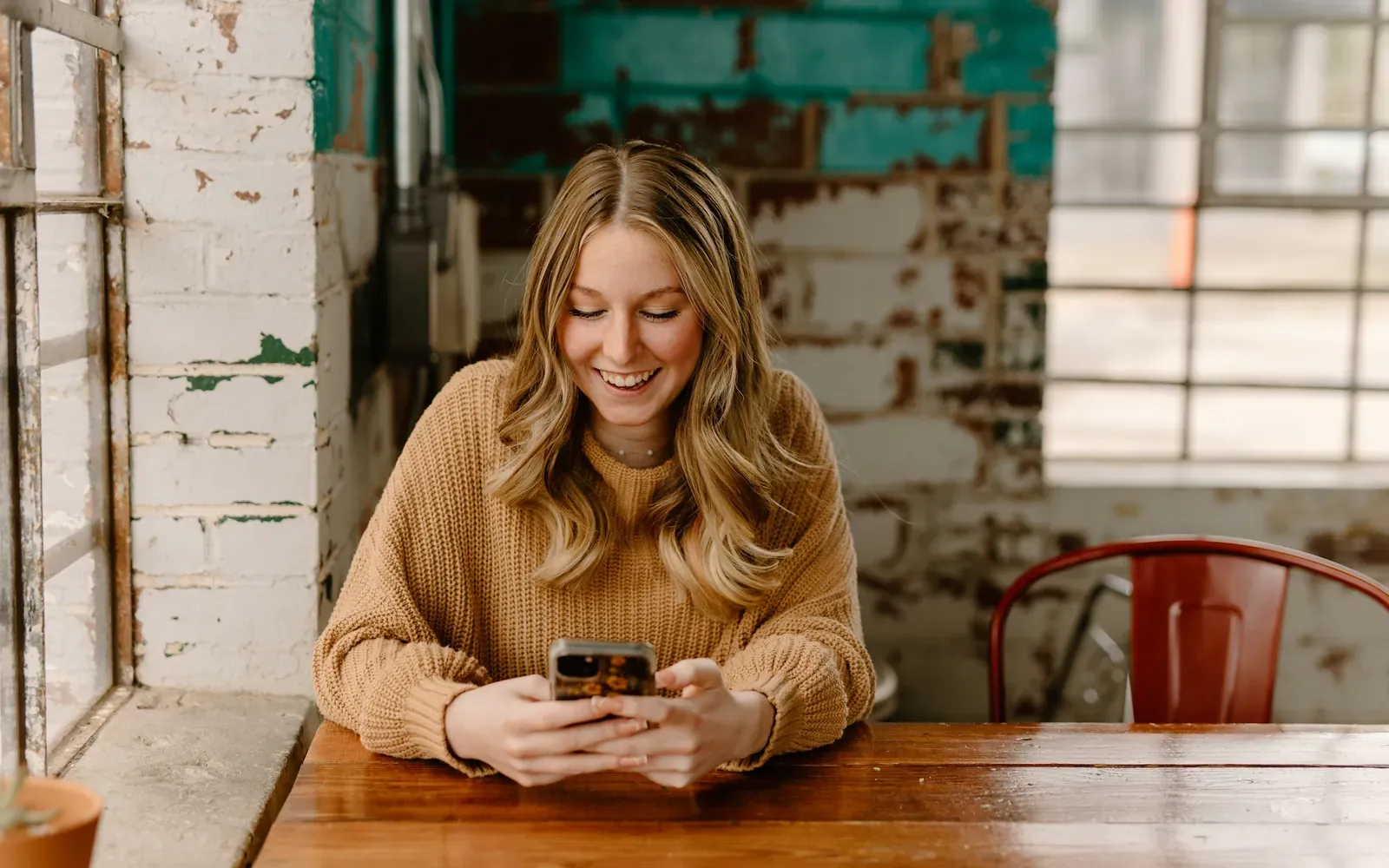 Smiling woman checking her phone at a wooden table, representing an applicant checking for a USCIS case status update.